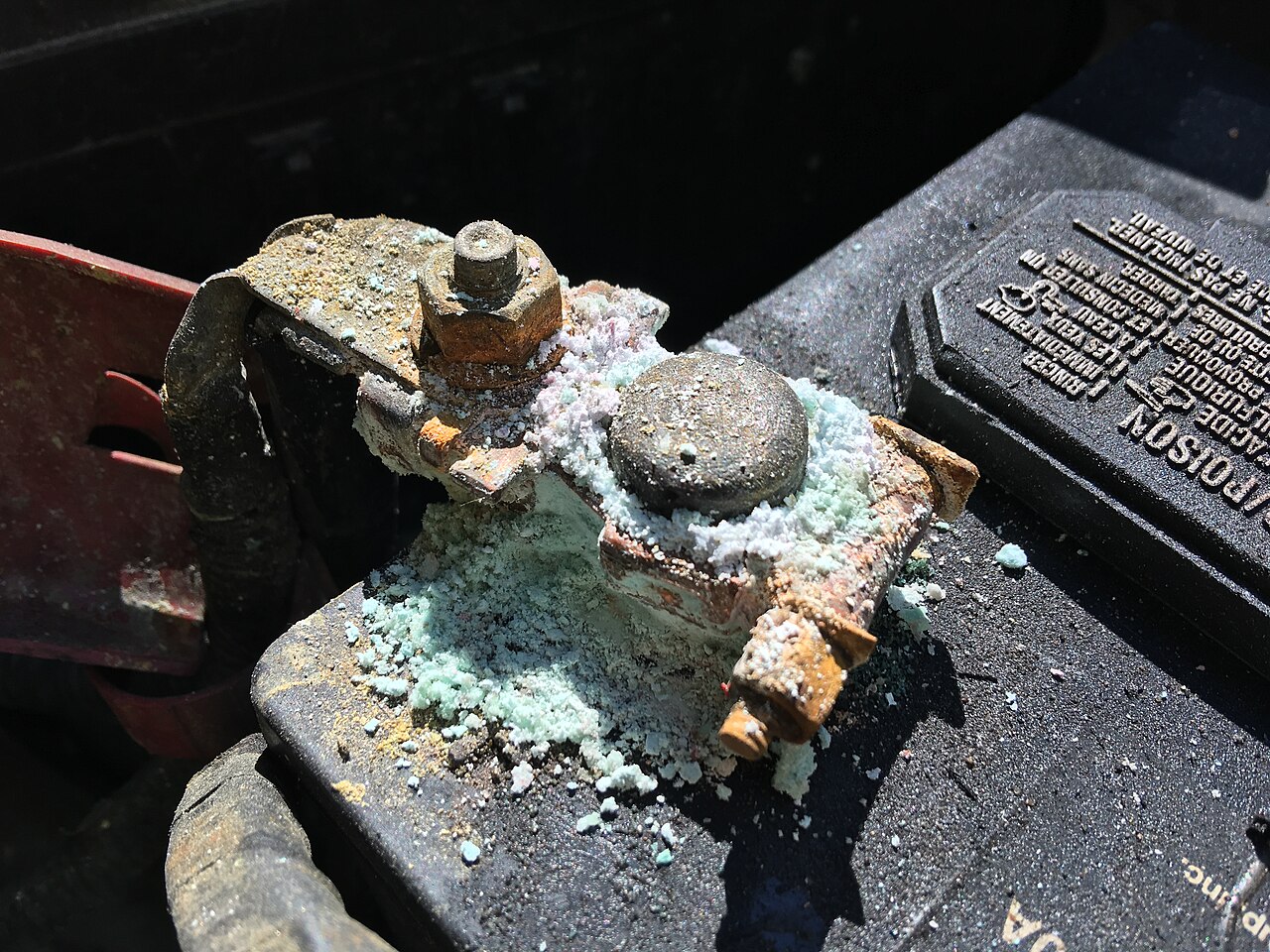 Close-up of a lead-acid battery terminal clamp buried in white and pale green crystalline corrosion, with a heavily rusted bolt and orange rust staining across the clamp and battery top.