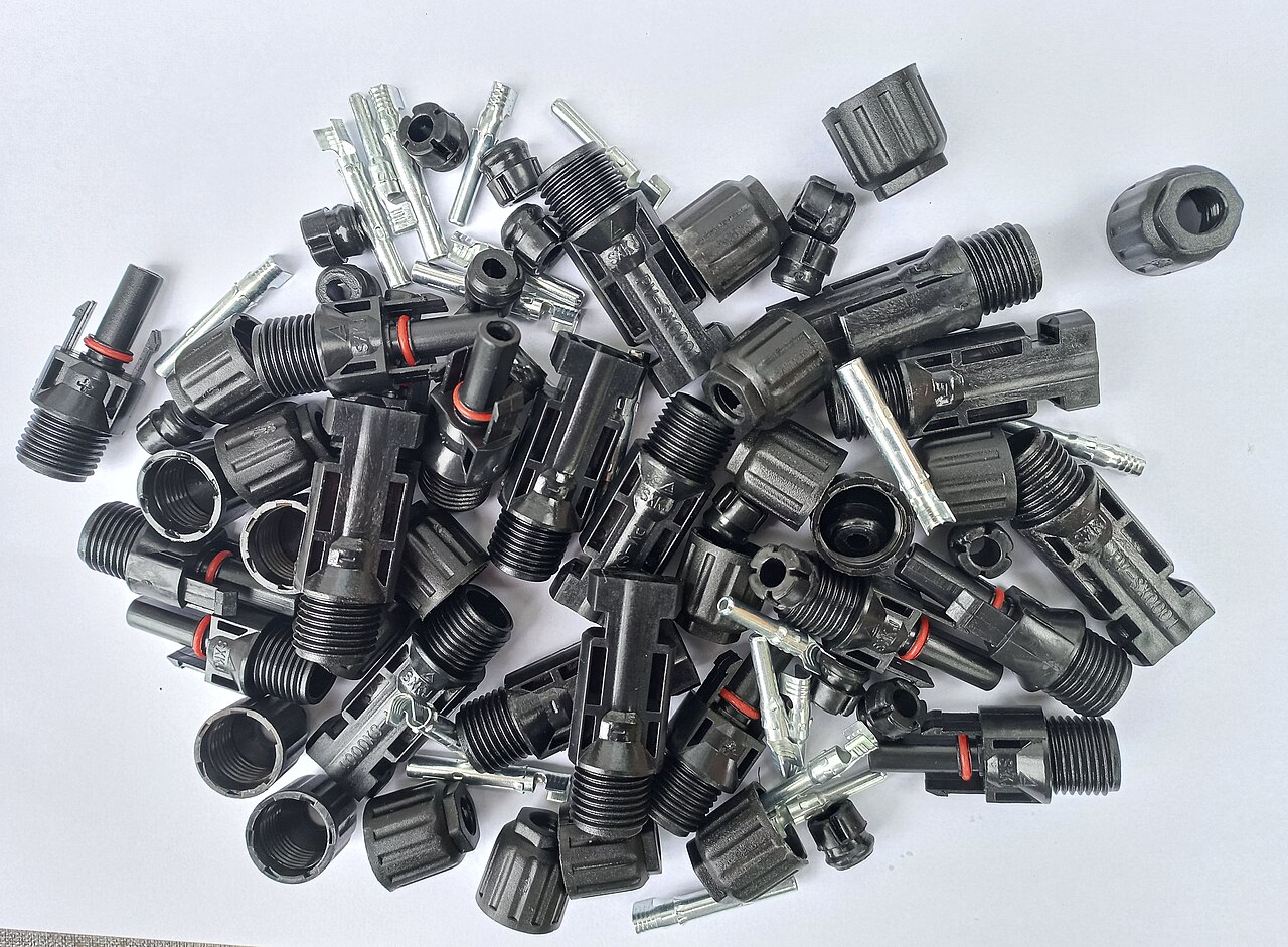 A pile of disassembled MC4 connector parts on a white background: threaded black plastic housings, ribbed gland nuts, red rubber sealing rings, and loose silver metal crimp contacts in both pin and socket form.